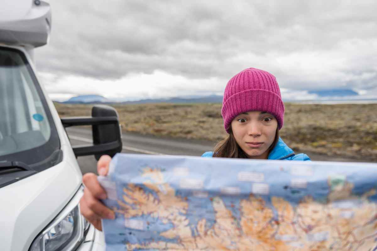 A tourist in Iceland wearing a fuchsia cap stares with an astonished face at a physical map of the country while her camper is parked on the road.