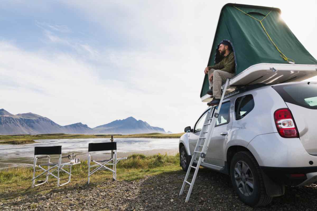 A man, from the top of his rooftop tent, looks out over the mountainous landscape surrounding him in Iceland while there are two folding chairs with a removable table and a thermos to his left, as well as a ladder to climb down from the top of the 4WD vehicle.