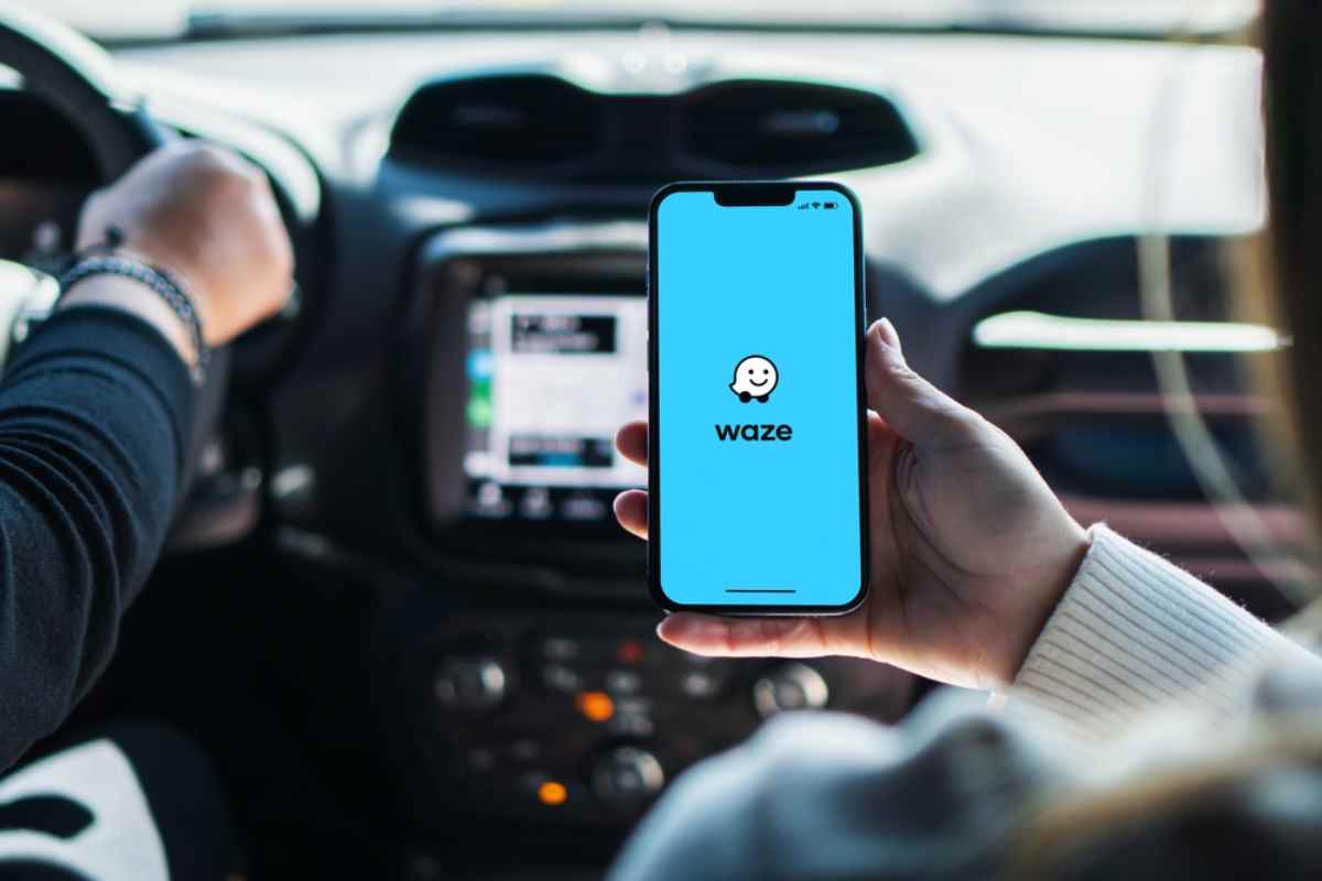 A hand holds a smartphone on which the Waze app logo is projected while next to it someone is driving a vehicle.