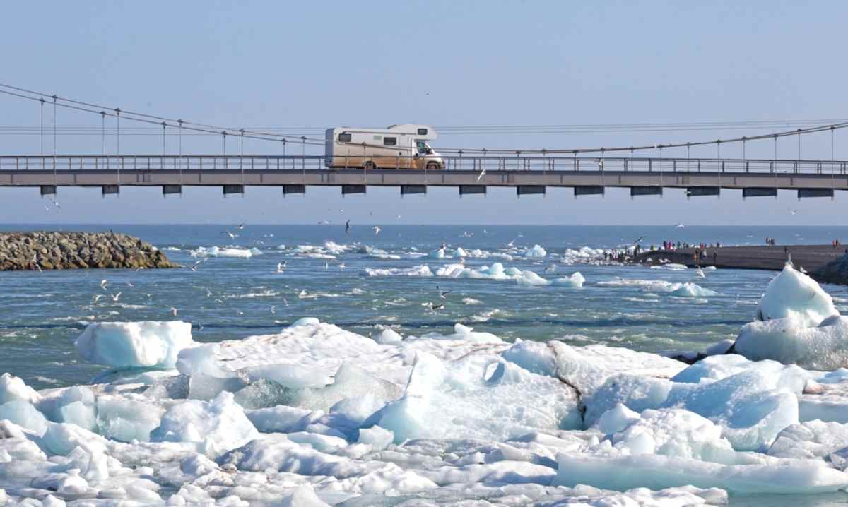A motorhome drives over a bridge in Iceland suspended in what looks like an open sea with large blocks of ice floating on it.
