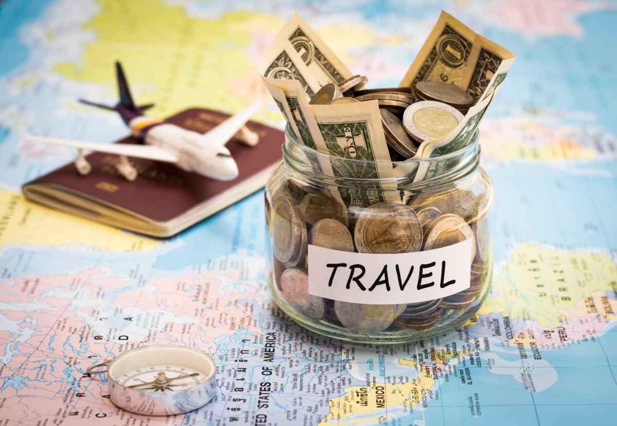 Glass “Travel” savings jar stuffed with cash and coins beside a passport and toy airplane on a world map.