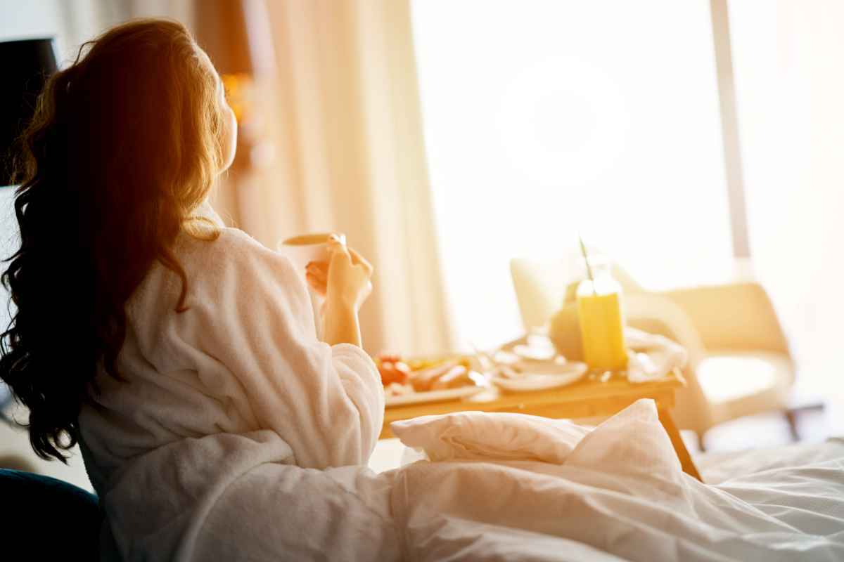 Woman in a hotel robe enjoying room-service breakfast by a bright window.