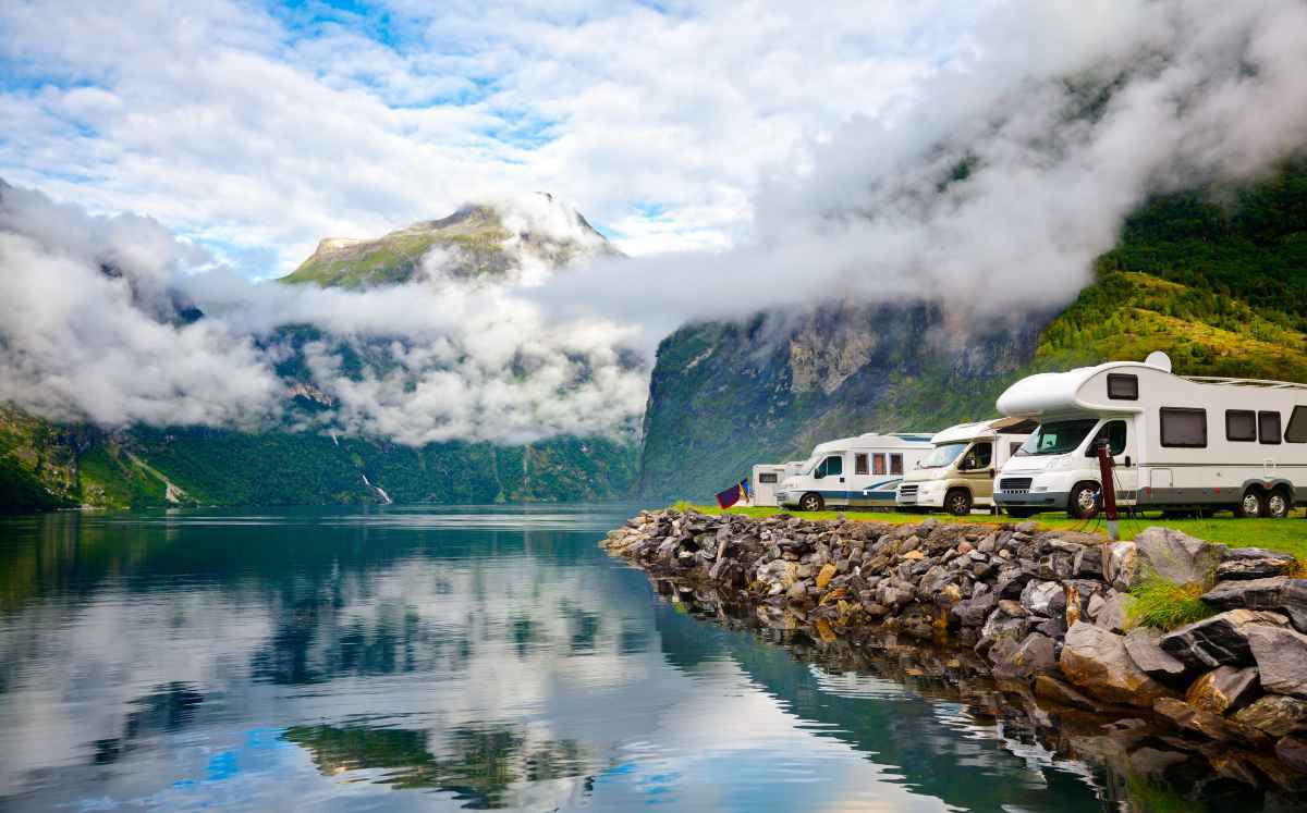 Row of RVs parked beside a mirror-calm fjord with misty mountains.