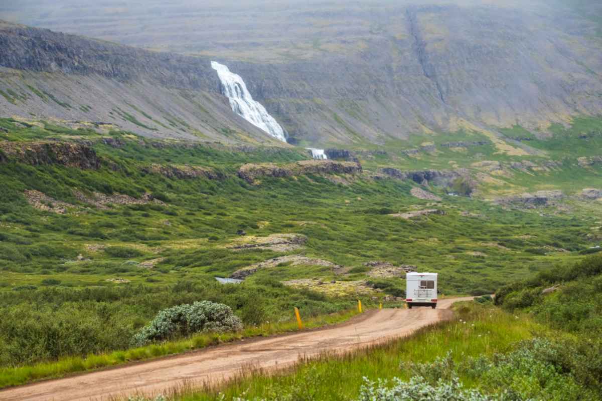 A white camper approaches an Icelandic waterfall in the Westfjords in the distance.