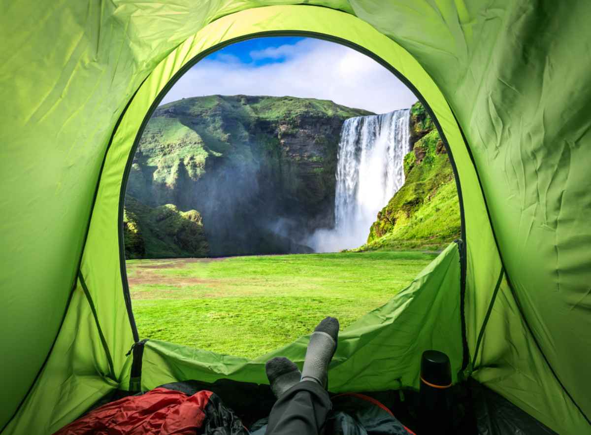 Feet peek through the opening of a tent near a spectacular Icelandic waterfall in a green meadow.