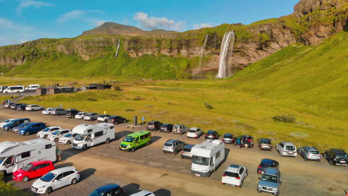 A large number of vehicles, including campers, park in front of an Icelandic waterfall located in the rural area.
