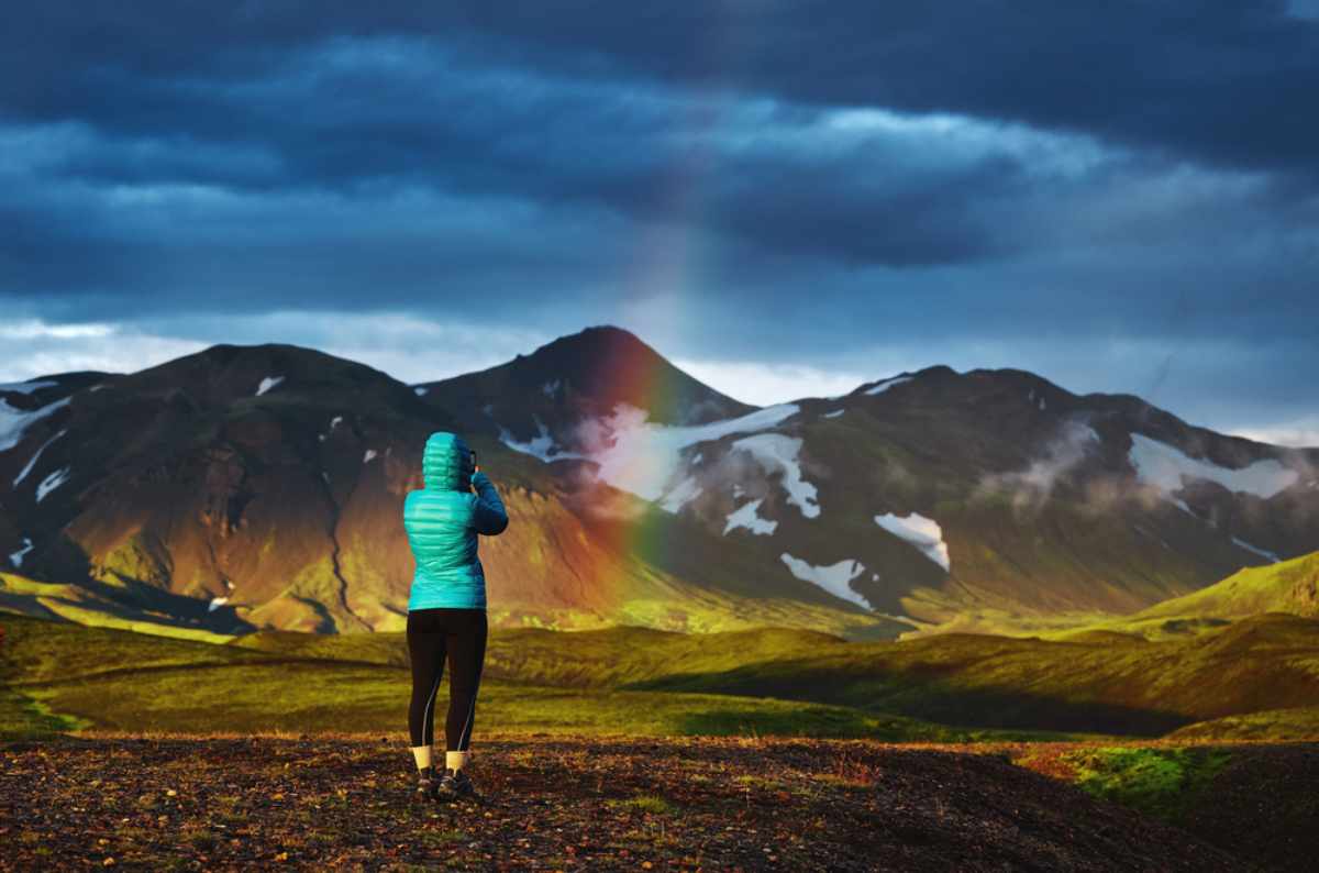 Person in a teal puffer jacket photographing a rainbow over Icelandic mountains at sunset.