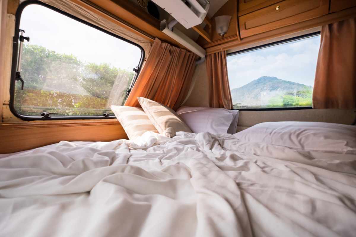 Cozy campervan bed with rumpled white linens and mountain views through side windows.