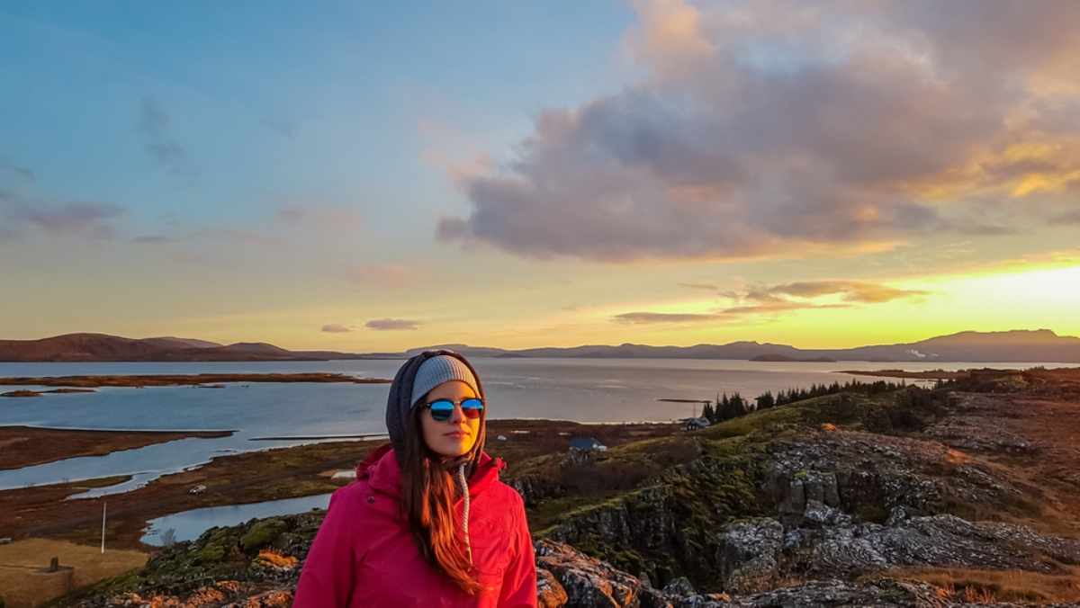 Woman in a beanie and sunglasses at golden hour, overlooking a calm coastal landscape.