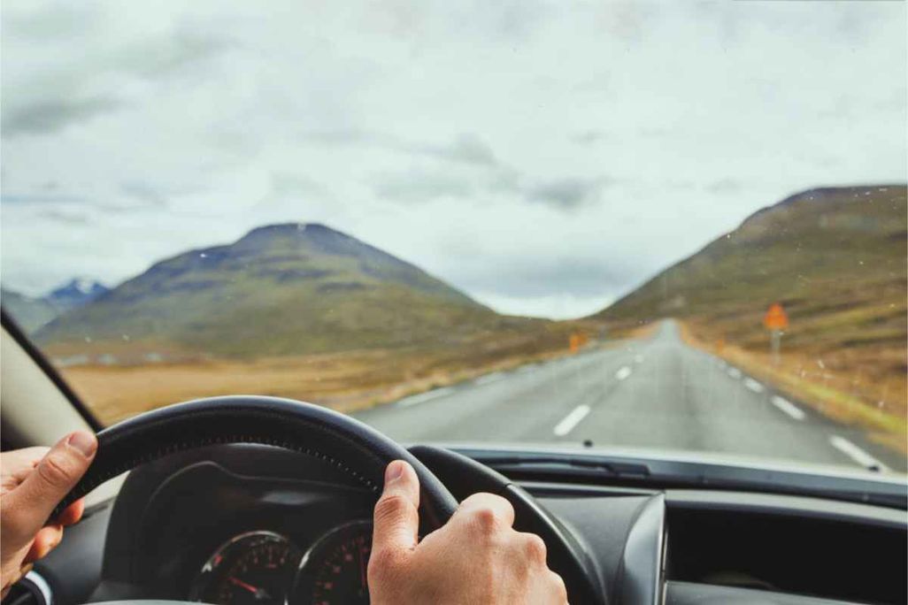 Hands on a steering wheel of a car driving down a paved road in Iceland, with mountains in the distance and cloudy skies overhead.