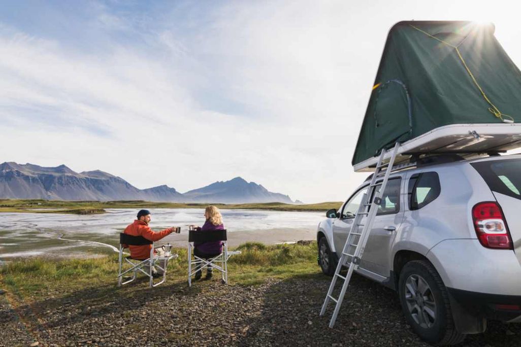 A couple sitting in camping chairs by a river, toasting with mugs next to a white SUV equipped with a roof tent, with mountains in the background.