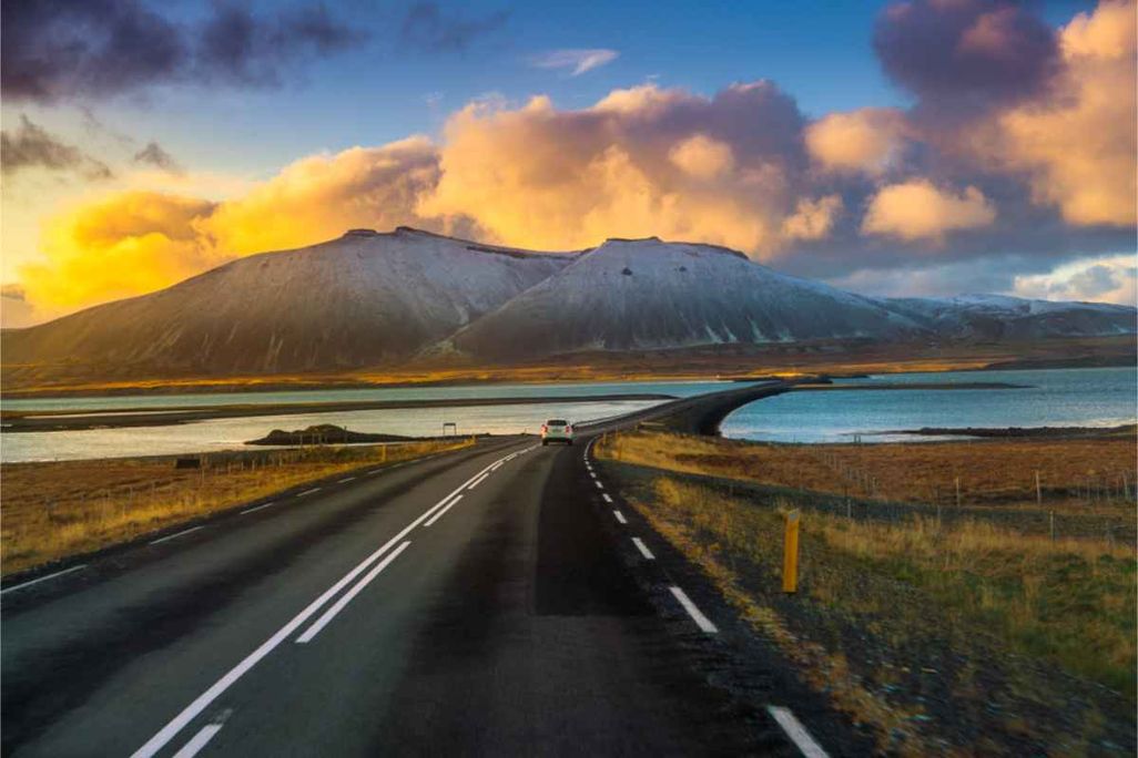 A car driving on a paved road over a bridge in Iceland, surrounded by water and mountains with snow-capped peaks under a colorful sunset sky.