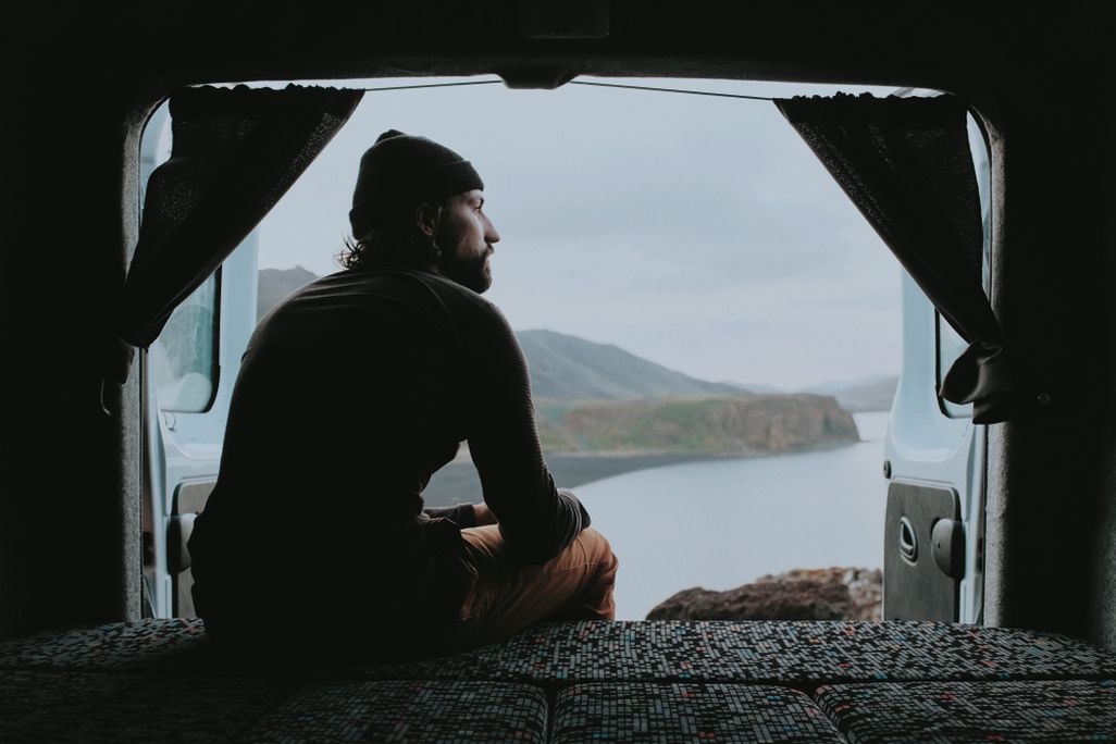 A man sitting at the back of a campervan, looking out over a calm river and distant mountains, framed by the van’s open rear doors and curtains.