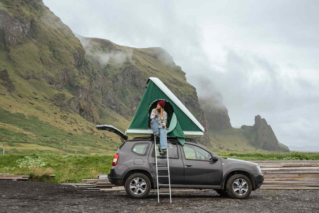 A woman sitting in a green roof tent mounted on a gray SUV, enjoying a drink with misty green cliffs in the background.