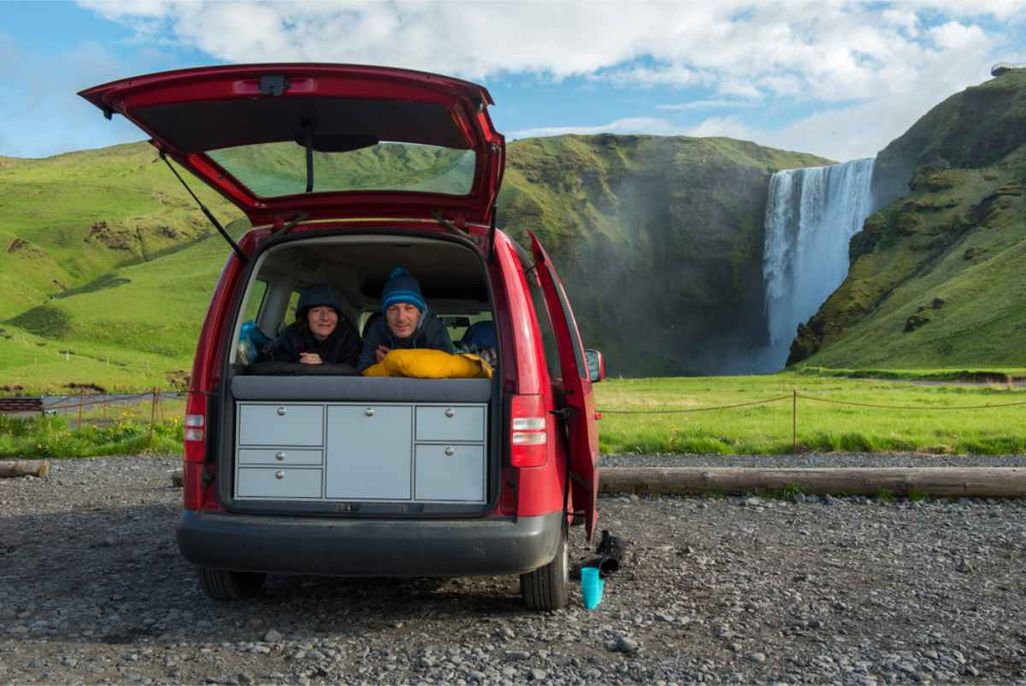 Two people lying in the back of a red campervan parked on gravel, looking out at the green landscape and Sk&oacute;gafoss waterfall in the background.