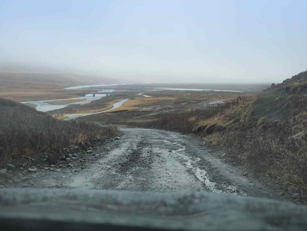 View from a vehicle driving on a rough gravel road in Iceland, with a river and flat plains in the distance under a foggy sky.