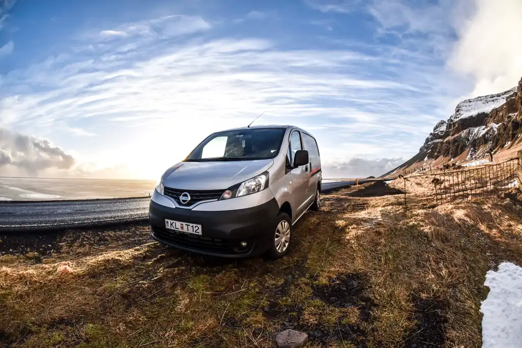A silver rental van parked on the side of a road in Iceland, with snow-dusted mountains in the background and sunlight breaking through partly cloudy skies.