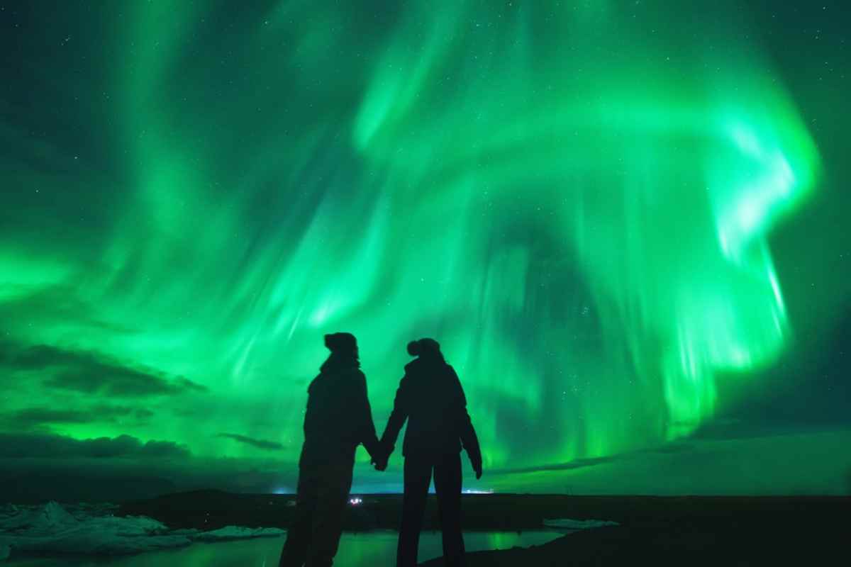 A couple, in the shade, enjoy the spectacular greenish Nordic lights in the Icelandic night sky head-on.