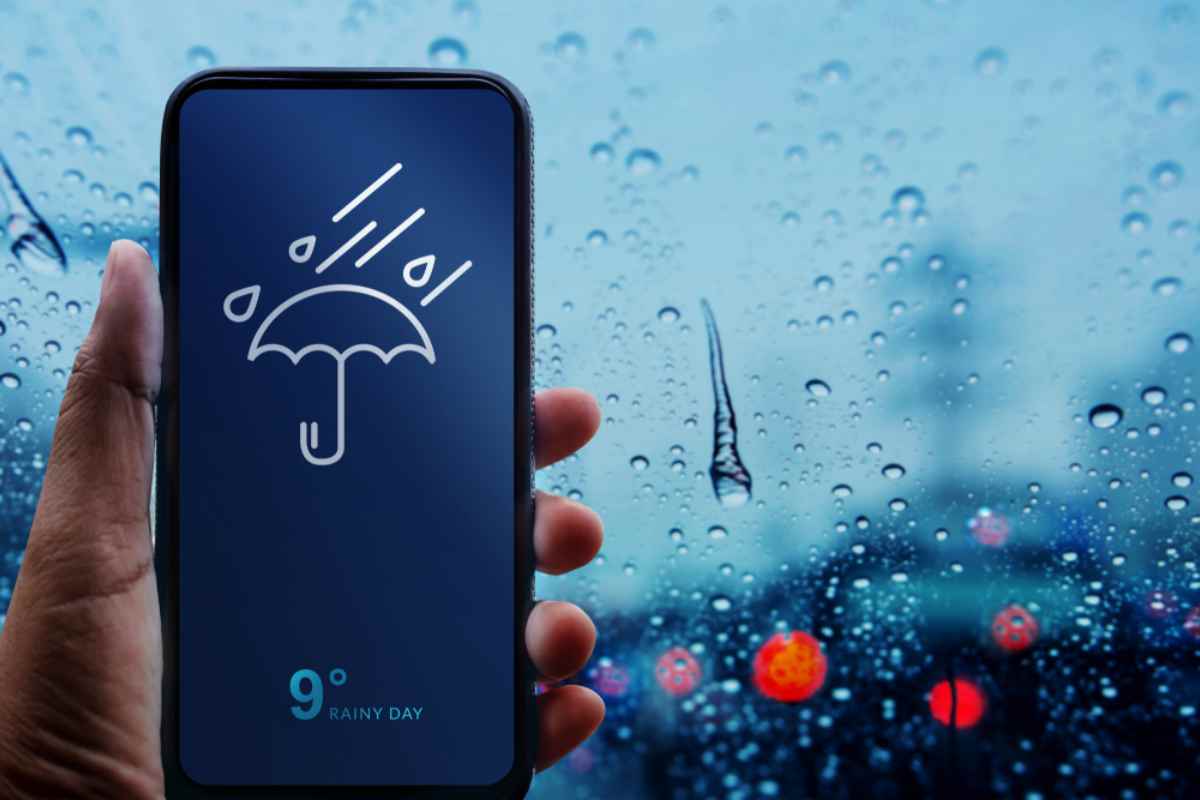 A hand holds a cell phone where a rainy weather forecast can be seen on the screen by the emoticon of an umbrella with drops that appears in the center. 