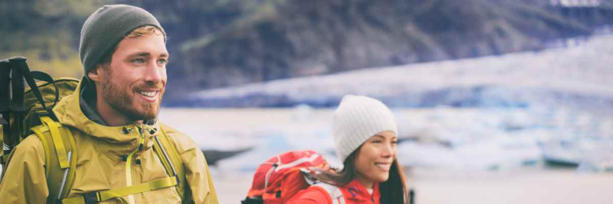 A couple, a boy in yellow and a girl in a wool cap and red, wear hiking clothes while walking near glaciers in Iceland.