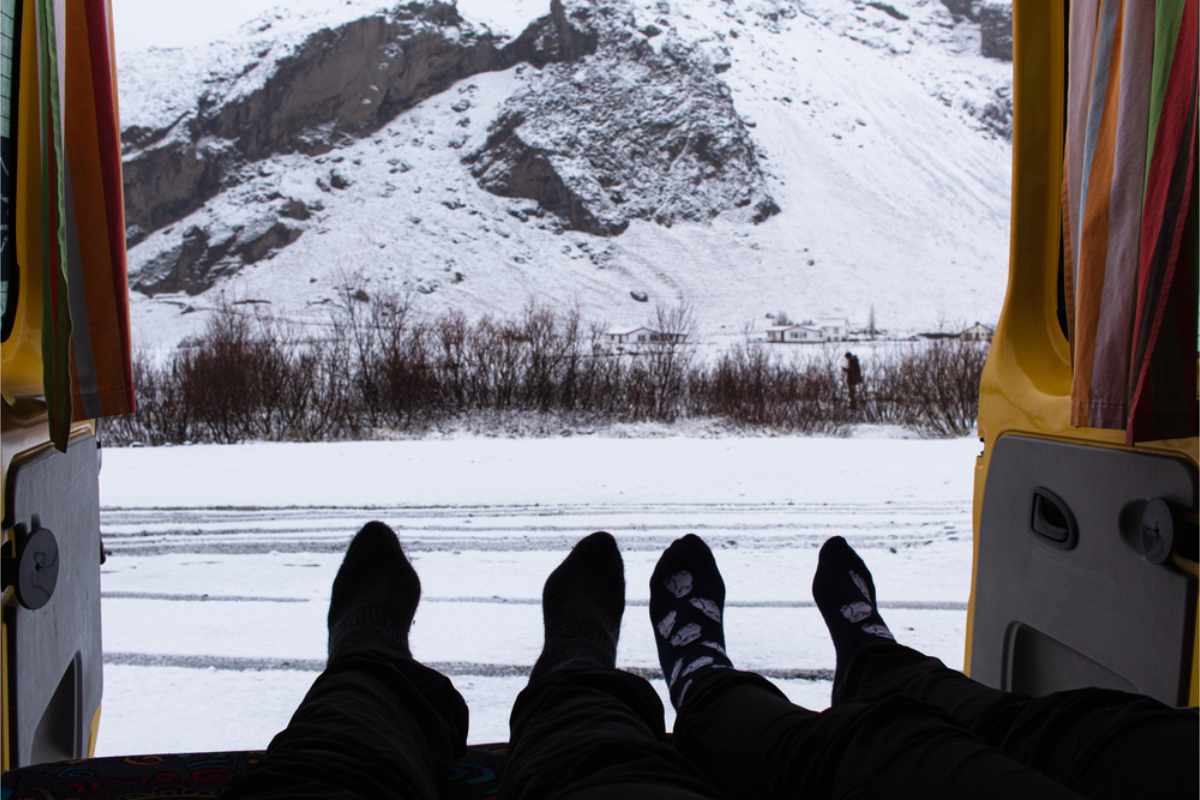 Two people relax in a van in Iceland with socked feet facing snowy mountains. A serene winter landscape is visible through the open doors.