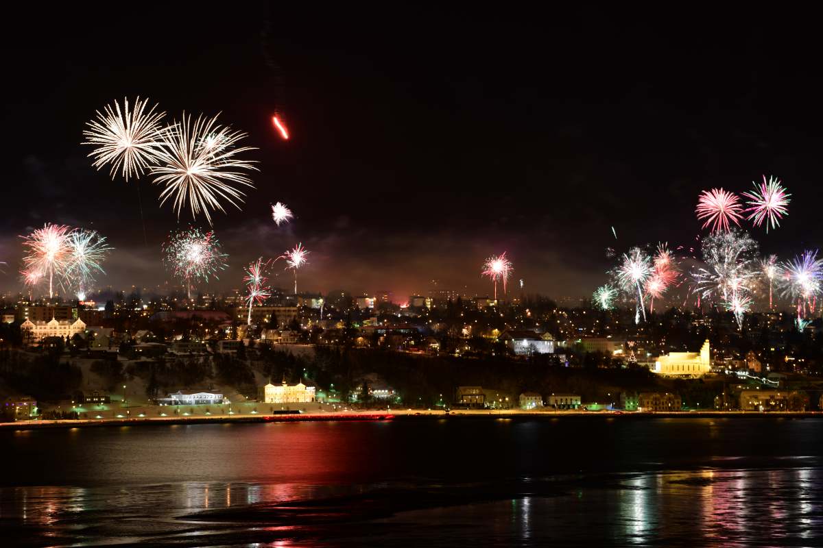 Fireworks light up the night sky over Reykjavik, reflecting on a river. Bright colors create a festive and celebratory mood.