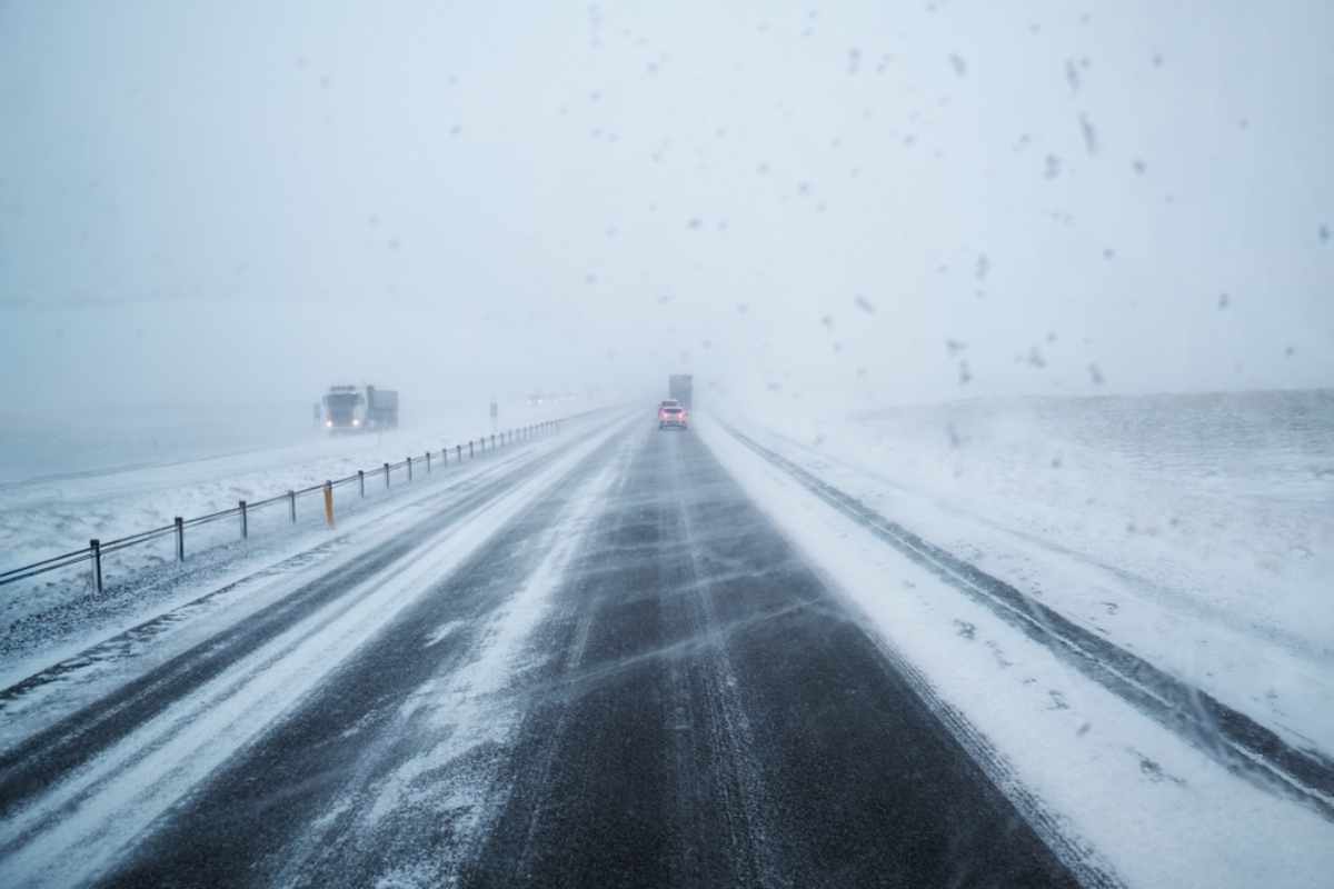 Snowy highway scene with cars and a truck driving in heavy snow. The road is partially covered in white, creating a bleak, wintry mood.