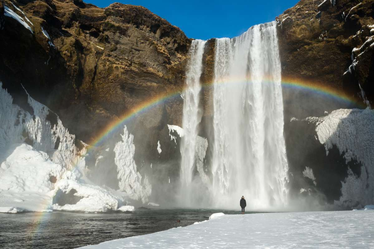 Person stands before skogafoss waterfall with a vivid rainbow arching in front. Snow and ice cover the ground, under a clear blue sky.