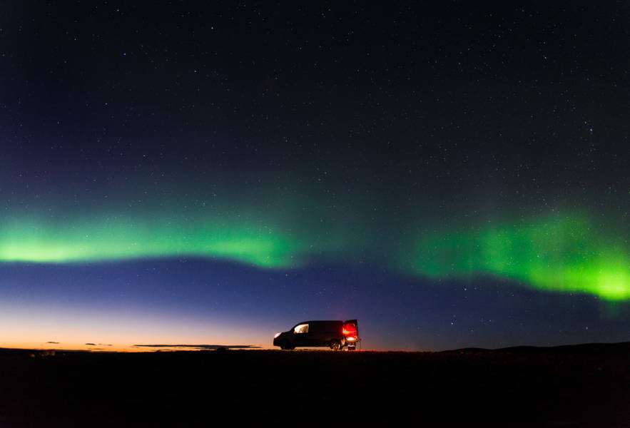 Van parked under vibrant green Northern Lights, vast starry sky above, creating a serene and awe-inspiring mood.