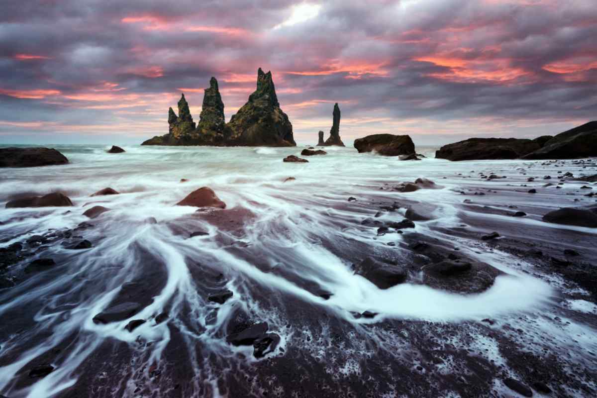 Dramatic seascape with towering rock formations against a vivid pink-orange sunset, swirling white waves on dark sandy shore. 