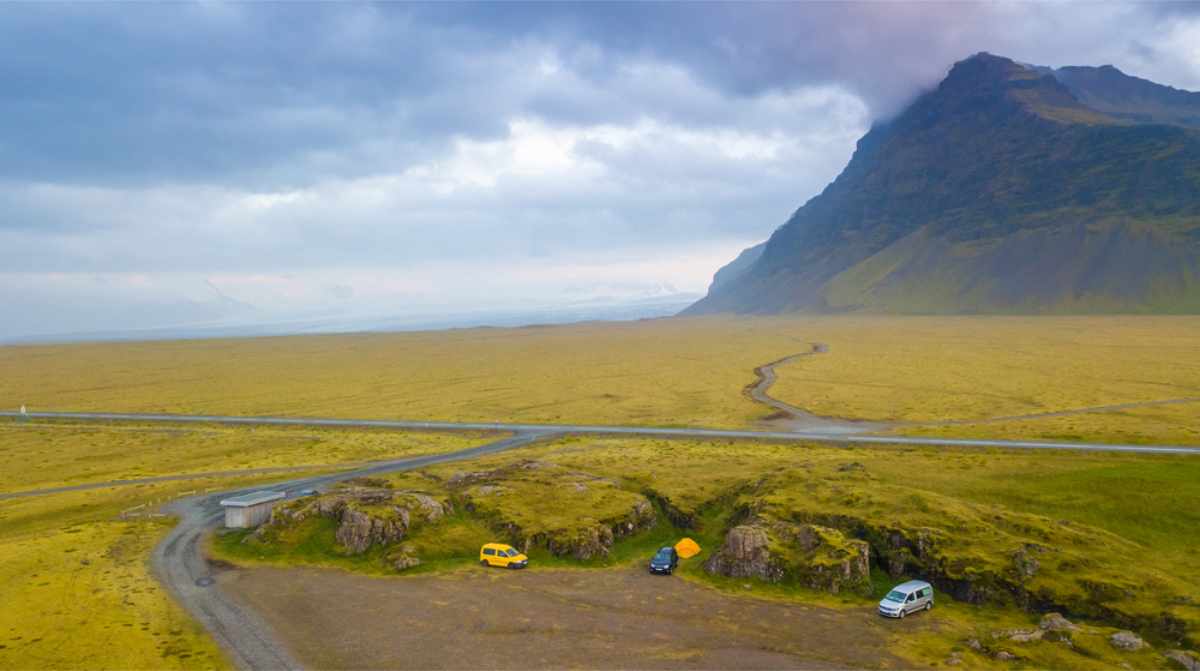 Yellow van and white car parked near a yellow tent in a vast, green field. A towering mountain under a cloudy sky dominates the background.