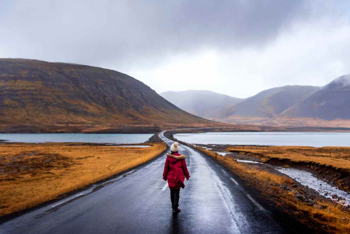 Person in red coat walks on a wet road between lakes and hills under a cloudy sky, evoking a calm and introspective mood.