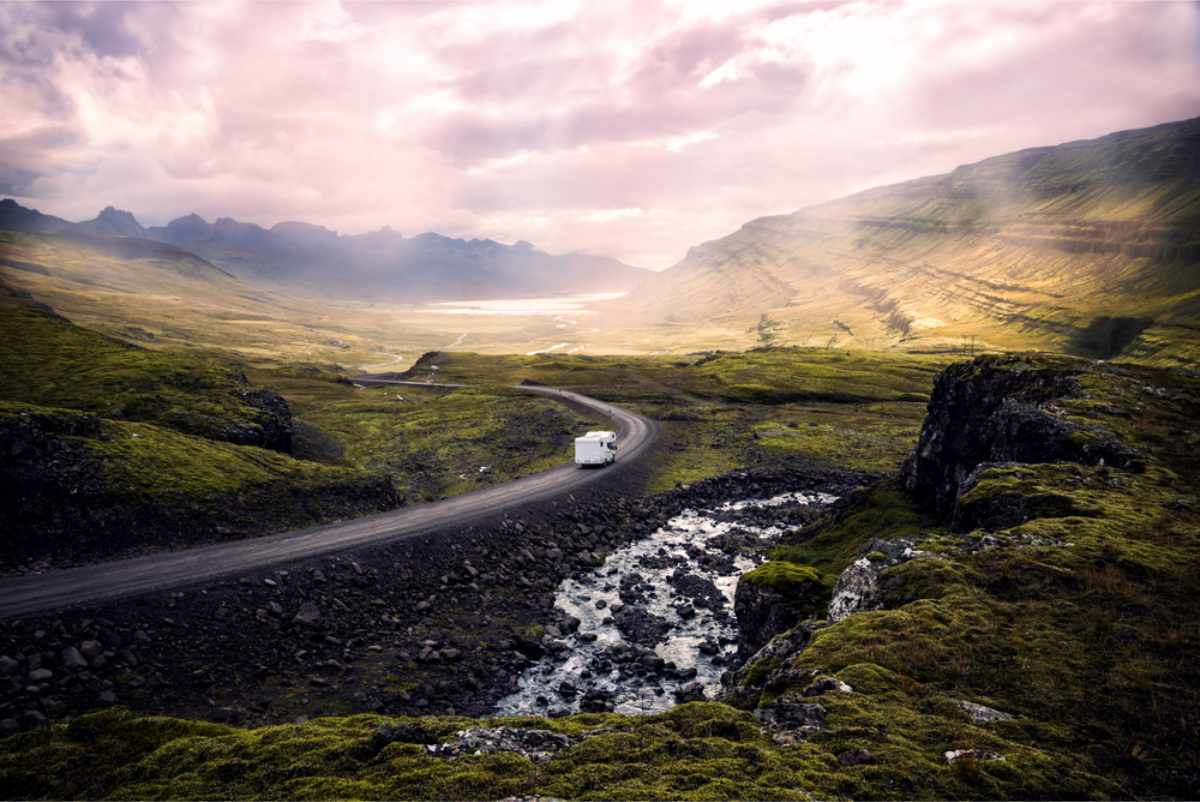 A white camper van drives along a winding road through a vast, green mountainous landscape under a cloudy sky with soft sunlight.