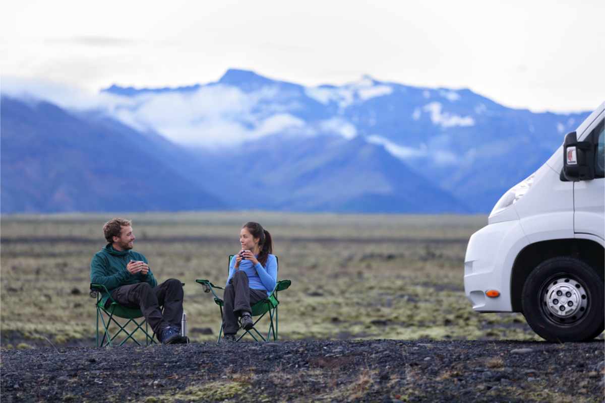 Two people sit on camping chairs, chatting with mugs in hand, in a vast field with a mountain backdrop. A white van is parked nearby.