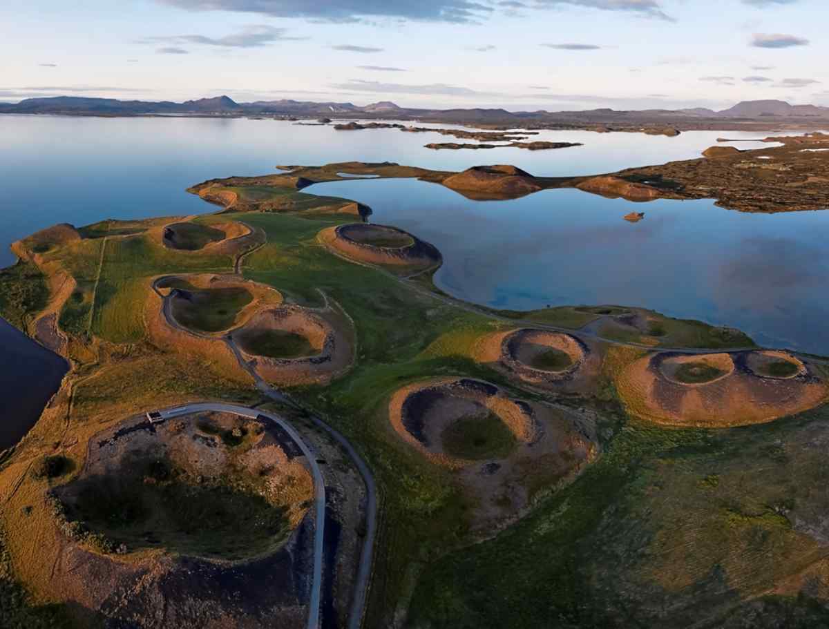 Aerial view of grassy pseudocraters in Iceland by a calm lake under a blue sky. The landscape features lush green and brown earth tones.