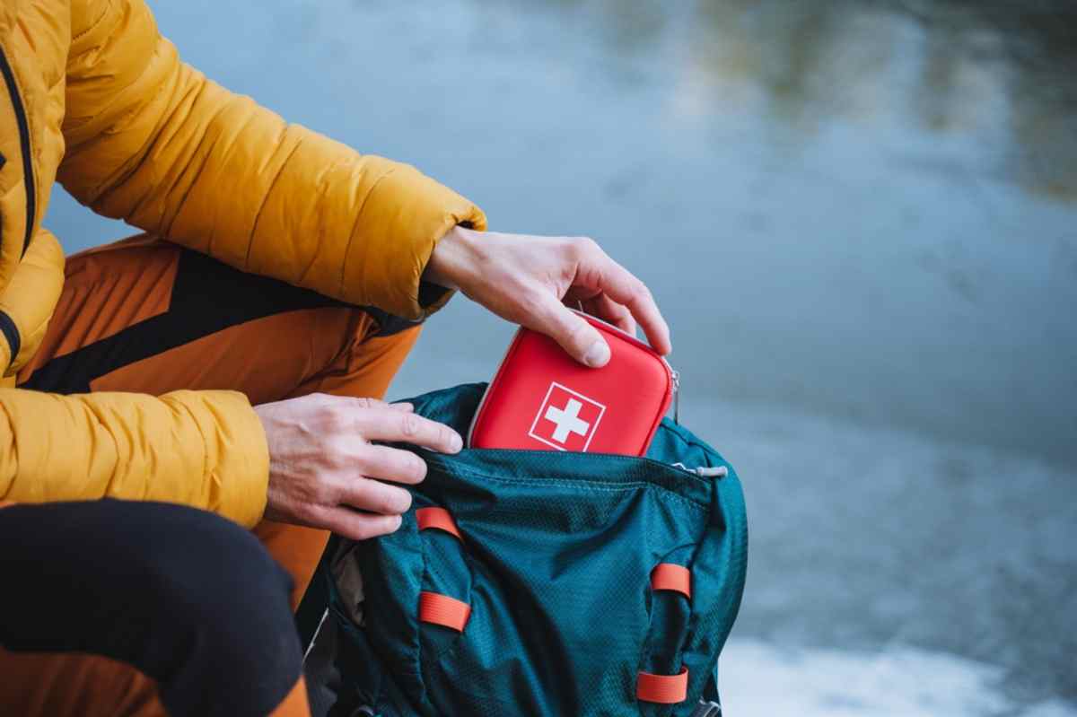 Person in yellow jacket places a red first aid kit into a blue-green backpack with orange straps, next to a calm body of water.
