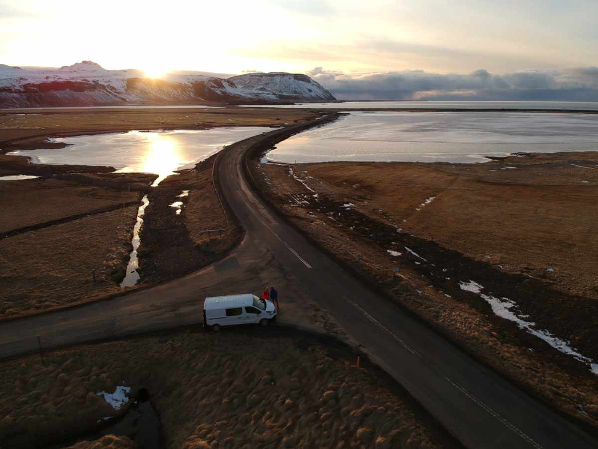Aerial view of a sunset over a snowy mountain with a winding road. A white van is parked on the roadside with two people nearby.