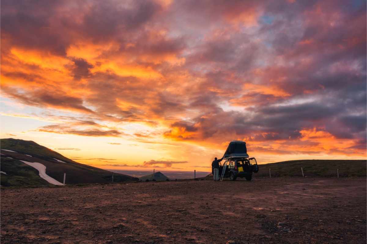 SUV with rooftop tent, person standing beside as vibrant orange sunset fills the sky. Rugged terrain and rolling hills in the background.