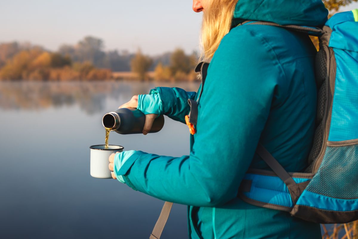 Woman in a teal jacket pours drink into a mug beside a serene lake. Background of blurred trees under a clear sky. Autumn setting.