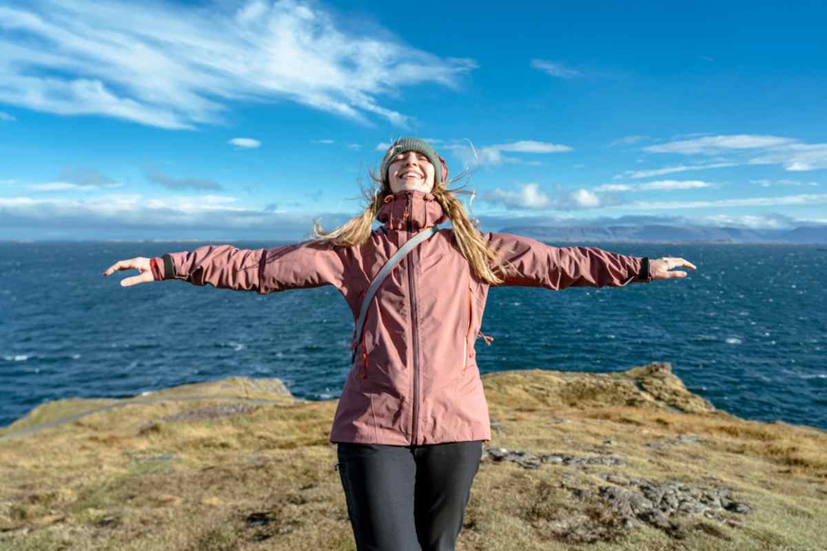 Woman joyfully extends arms on a cliff with ocean and blue sky in the background. She's wearing a pink jacket and gray beanie.