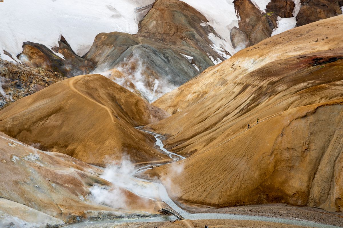 Steaming geothermal valley with orange-brown hills, snowy peaks, and a winding path. Two hikers ascend the trail, creating a serene mood.