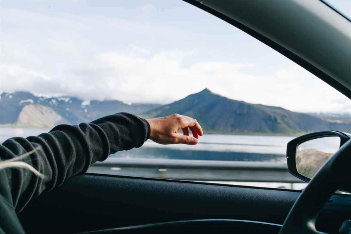 Hand in a black sleeve rests out car window, scenic mountain view, cloudy sky in the background. Calm and relaxed mood.
