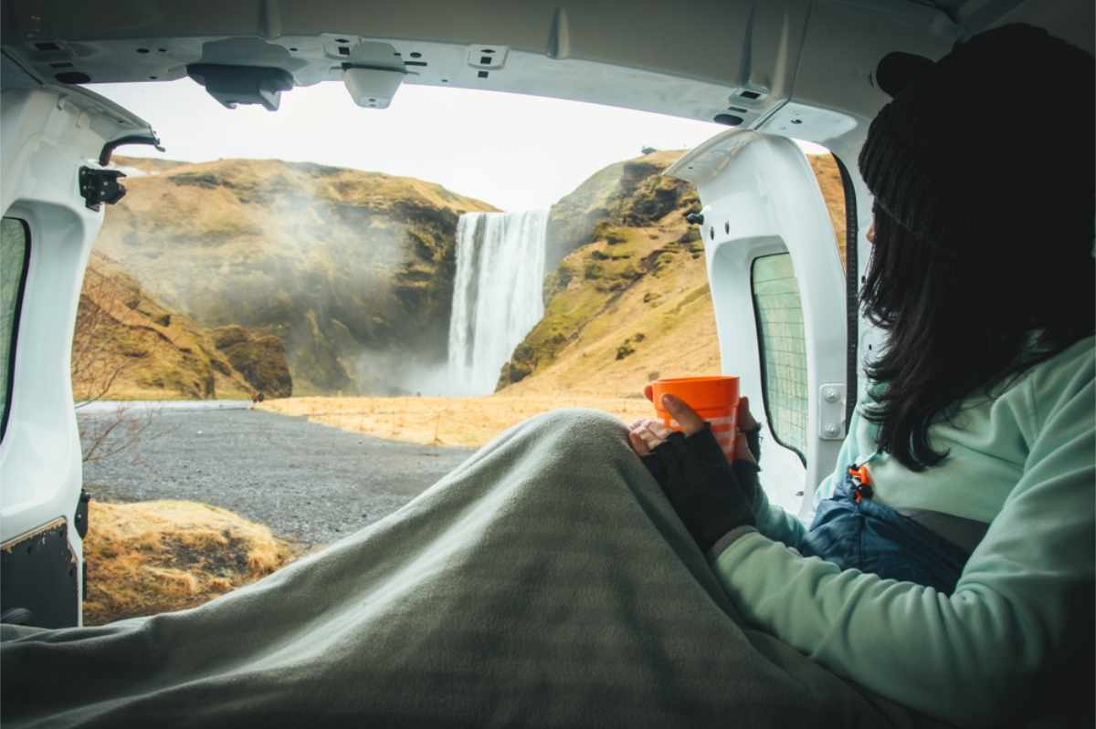 Person in a van holding an orange mug, wrapped in a blanket, gazing at Skogafoss waterfall. 