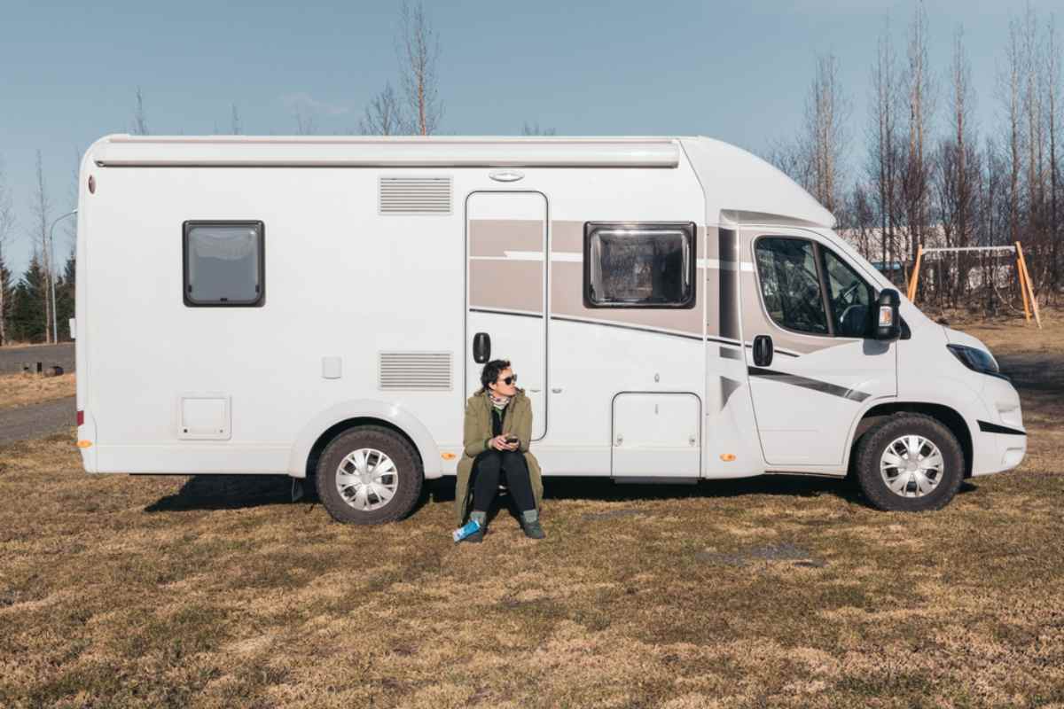 A person sits in front of a white camper parked on a clear day in a barren spot.