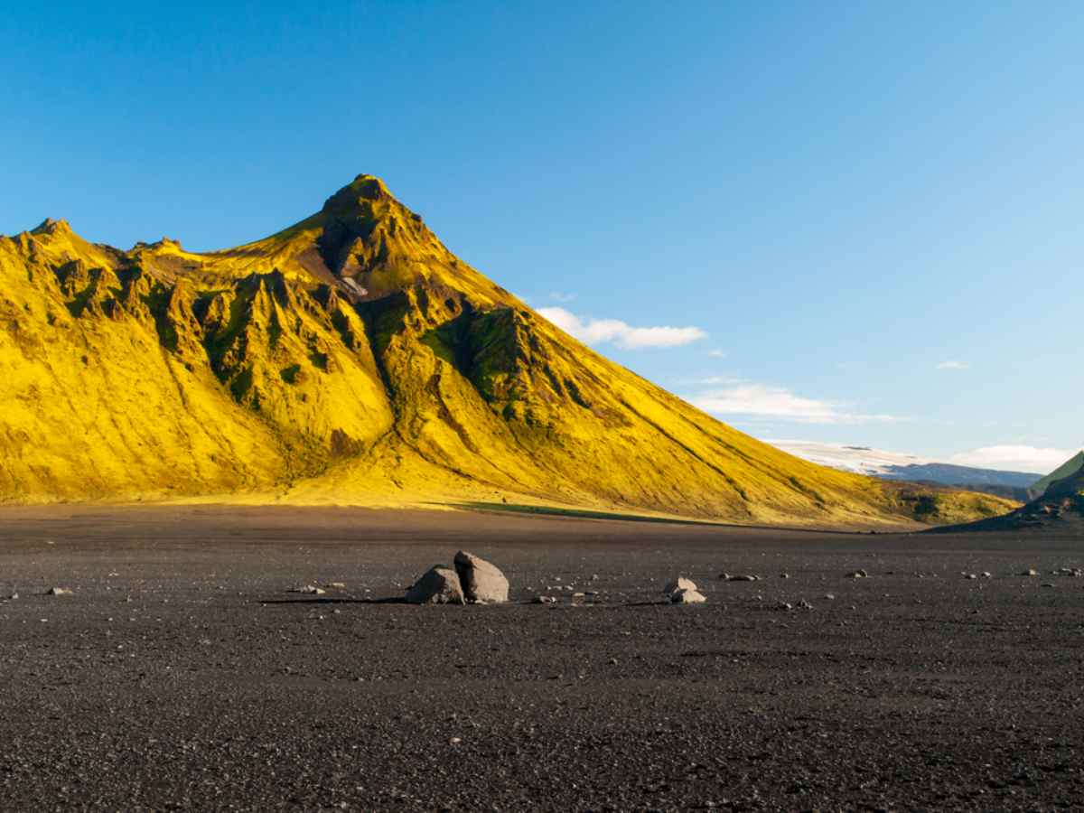 A steep mountain in Iceland appears completely bathed in sunlight next to a black and rocky landscape.