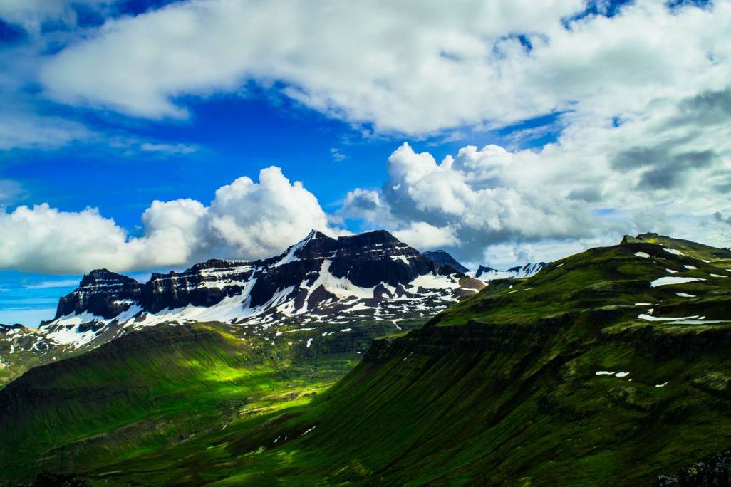Sunlit green valleys stretch toward rugged, snow-capped mountains under a partly cloudy blue sky in Iceland, highlighting the dramatic natural landscape.