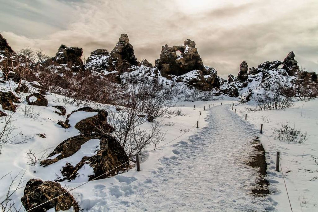 A narrow walking path covered in snow winds through dark volcanic rock formations in Iceland, with sparse vegetation and a cloudy sky above.