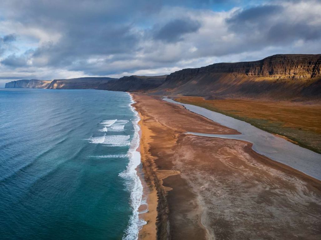A wide sandy beach in Iceland with gentle ocean waves on the left, a river running parallel on the right, and steep cliffs in the background under a cloudy sky.