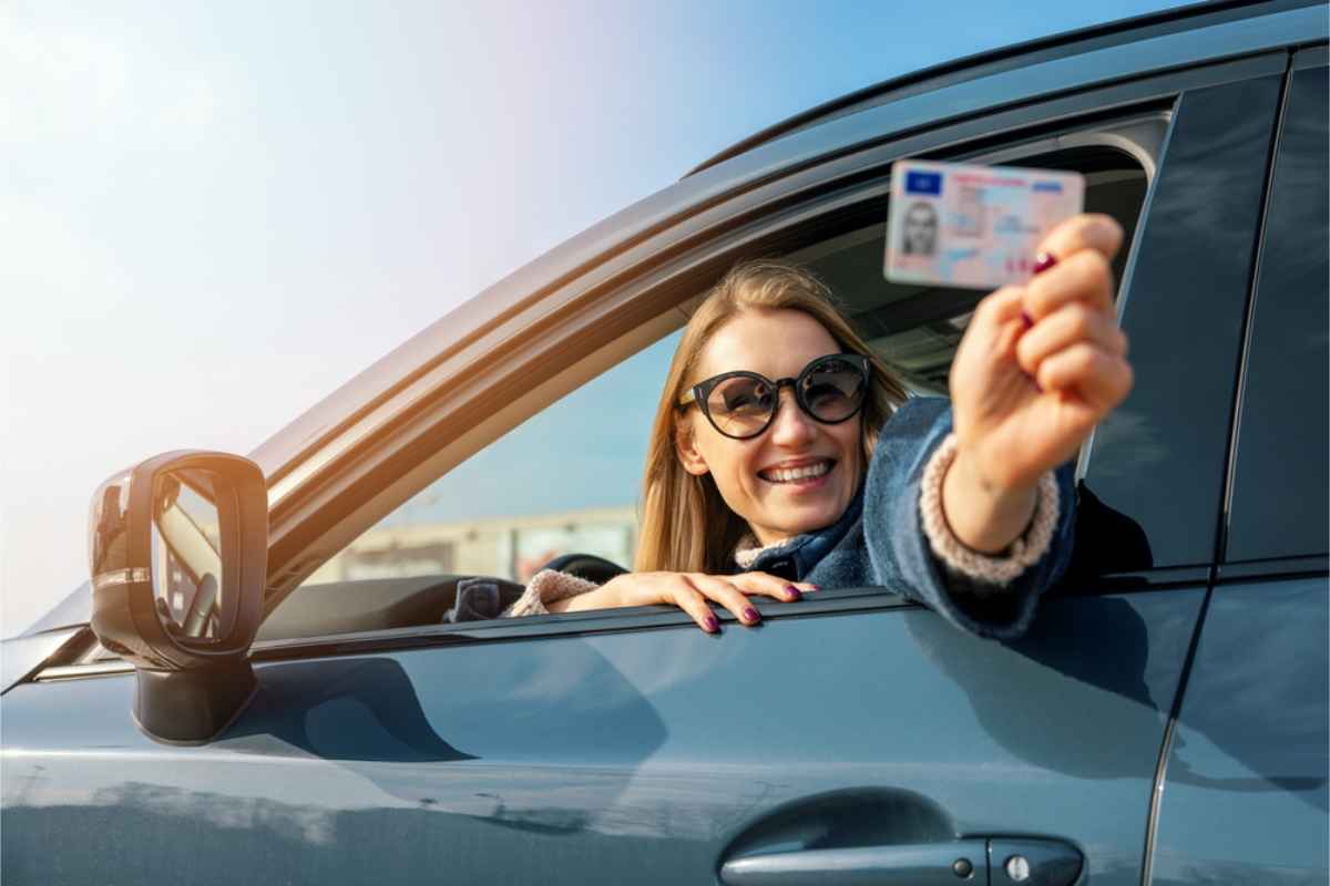 Smiling woman in sunglasses leans from car window, proudly displaying a driver's license, under a clear blue sky.
