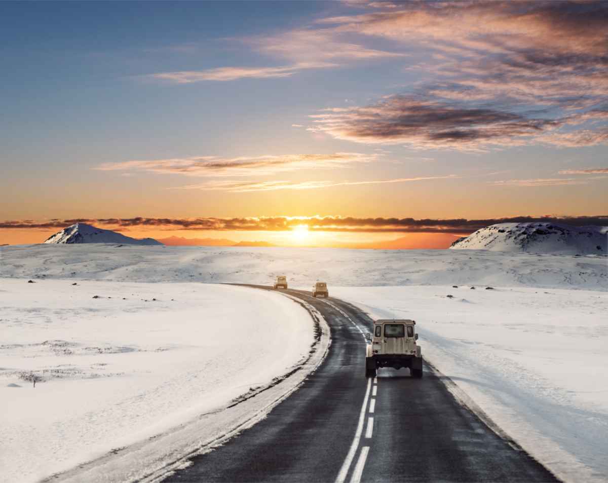 Vehicles on a snowy road at sunrise, surrounded by mountains and a colorful sky, creating a serene and adventurous atmosphere.